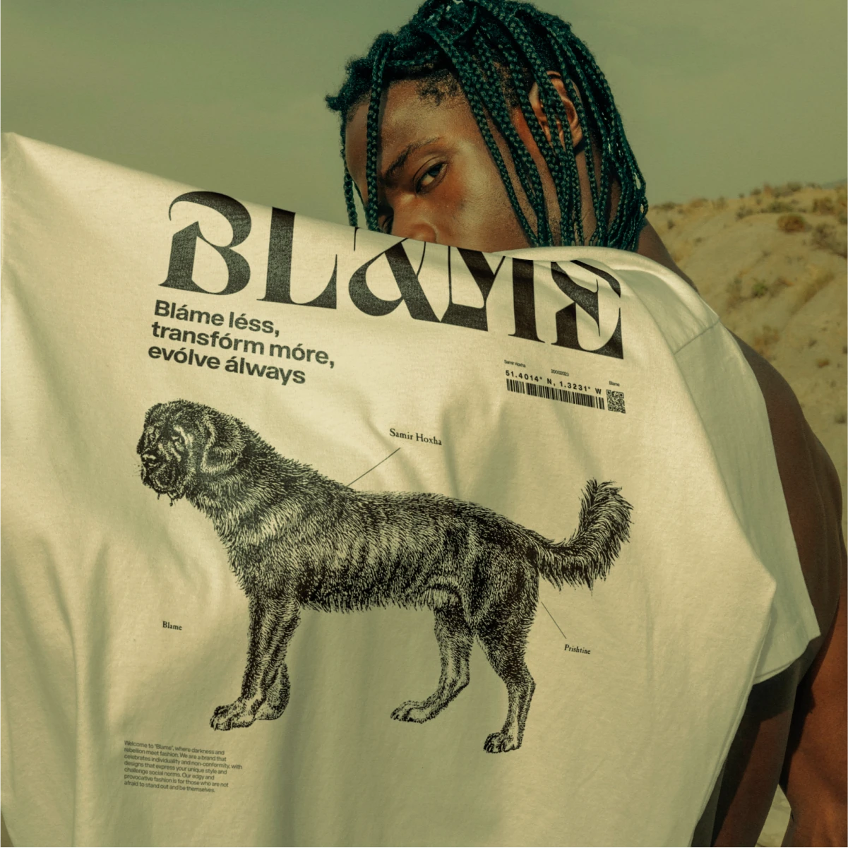 Man holding up a cream Blame t-shirt printed with the wordmark and an engraved dog, in the desert